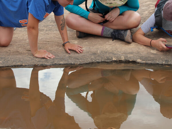 Three students observing a small pool of water in a nature, with their reflections visible